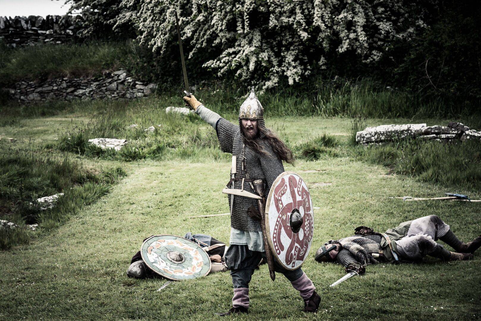 A re-enactor dressed in chainmail with a shield is victorious over warriors lying prone on the ground, The Whithorn Trust