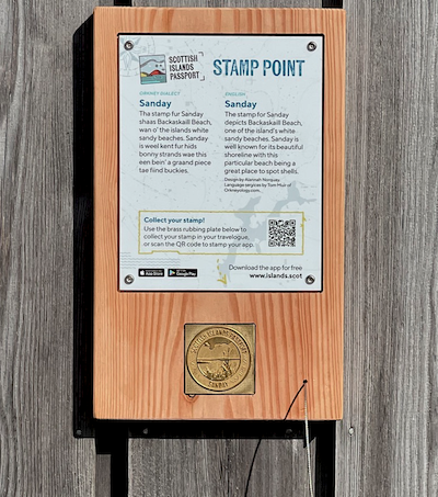 Photograph of a  Scottish Islands Passport stamping point showing an information board and a brass rubbing plate mounted on a wooden back