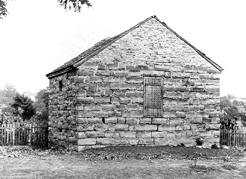 The Liberty Jail in an old, black-and-white photo. The walls are made of large stone bricks, and a wooden door sits in the wall a few feet above the ground.
