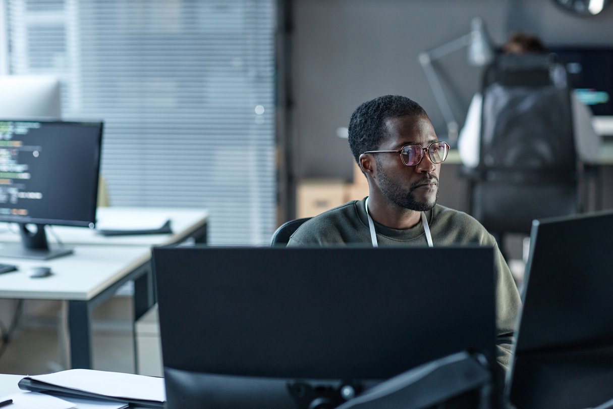 Professional wearing glasses works at a computer workstation in an office, focusing on multiple monitors while coding or reviewing information on screen.