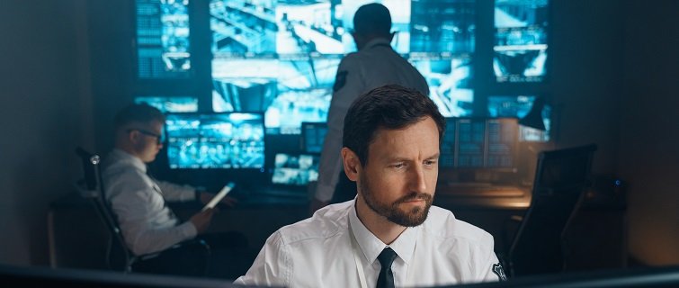 Businessman sits at desk looking at computer while behind him are video surveillance screens.
