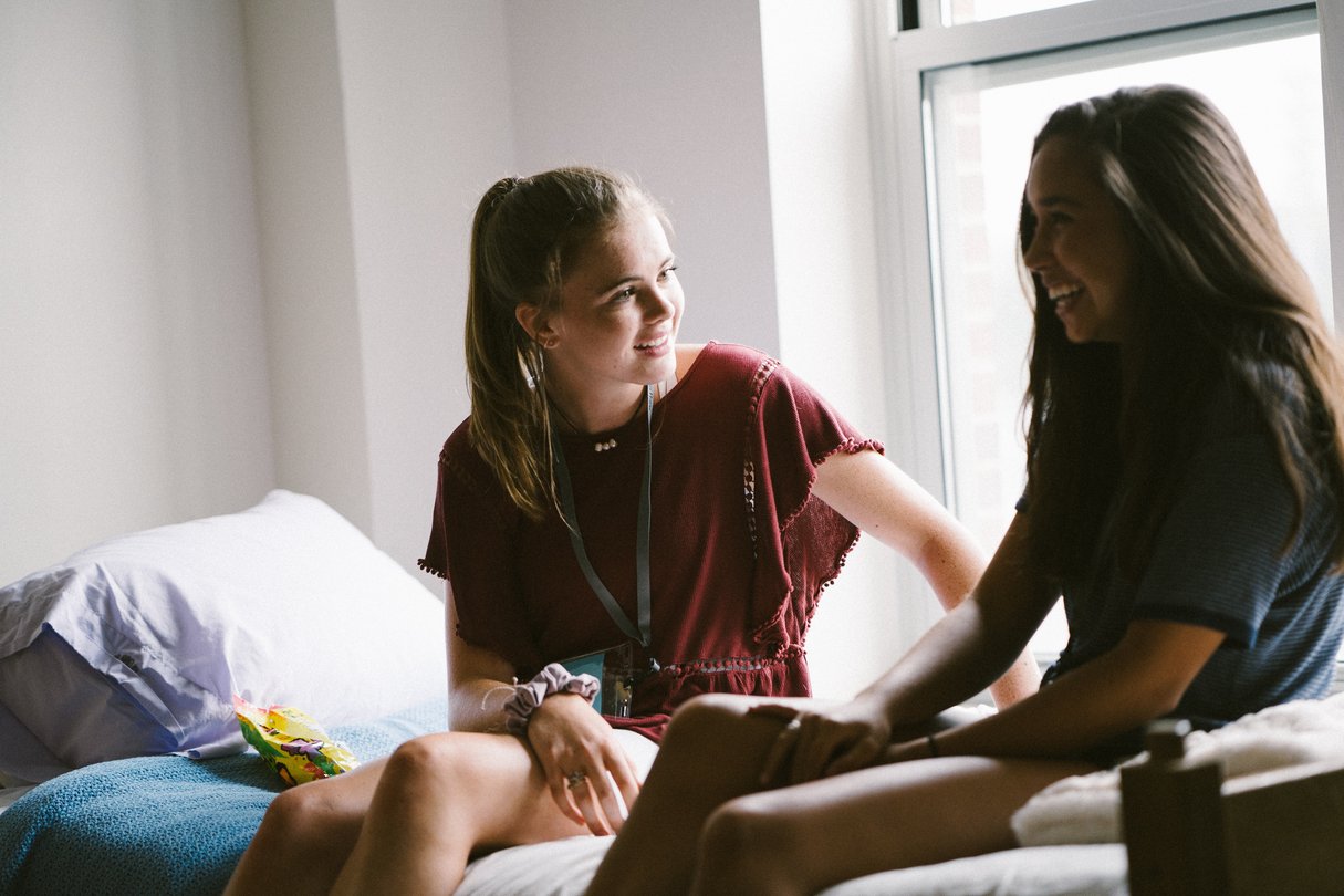 Two female students sitting in dorm bed laughing.