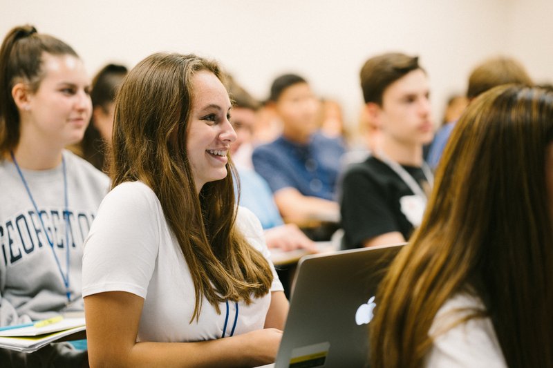 Students attentively listening in a classroom, one smiling while taking notes on a laptop.