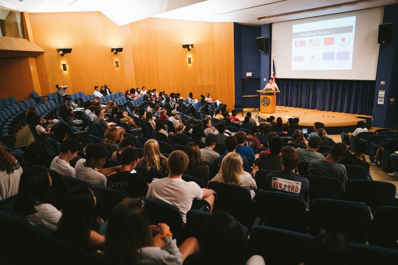 Students seated in an auditorium listening to a speaker presenting slides at a lecture.