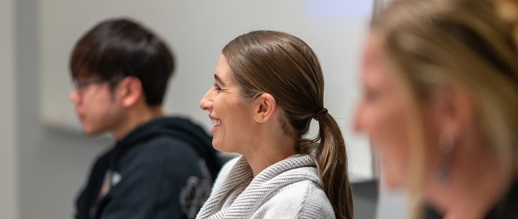 Students in modern classroom session. A woman in the center smiles while listening.