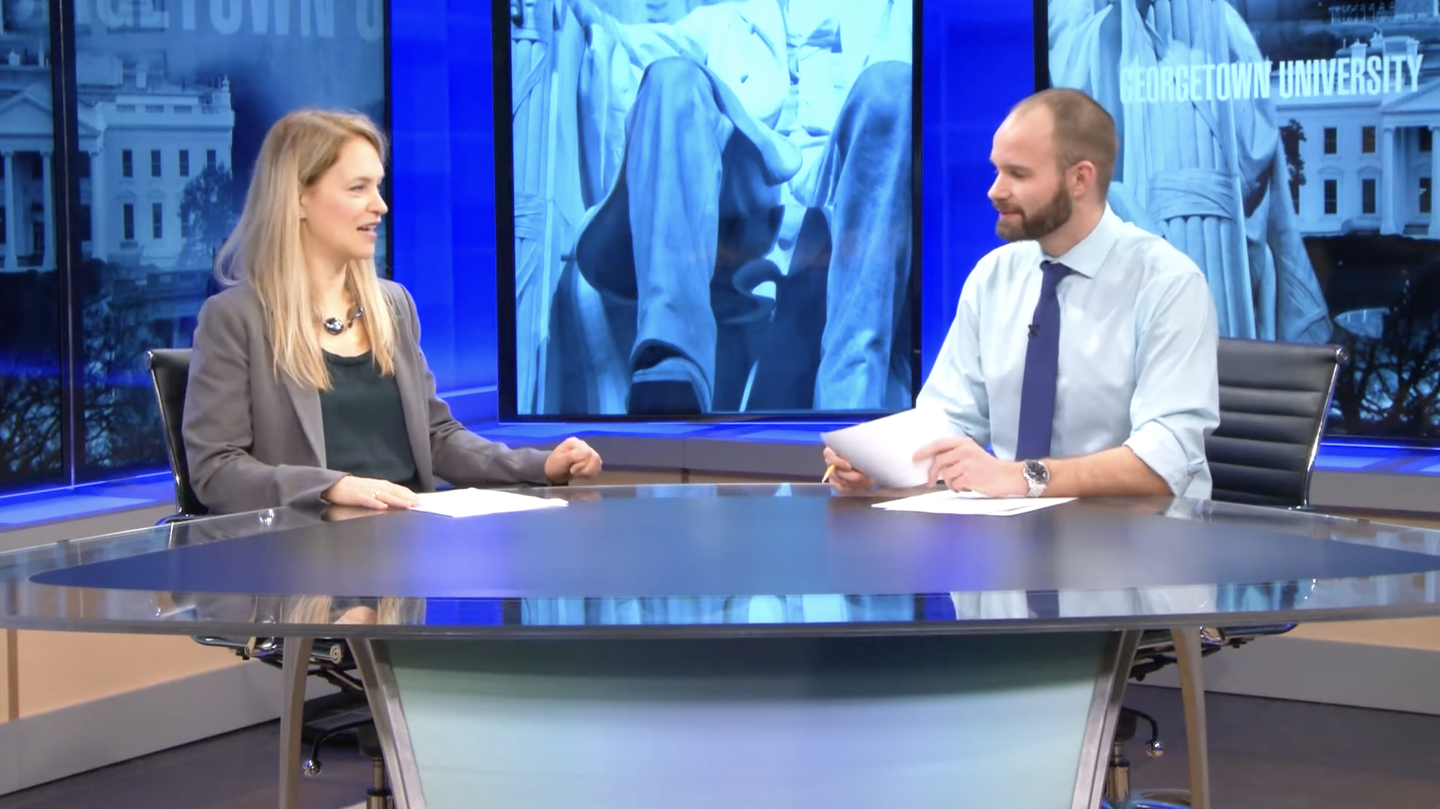 Two people sit across from each other at a studio news desk during a discussion.