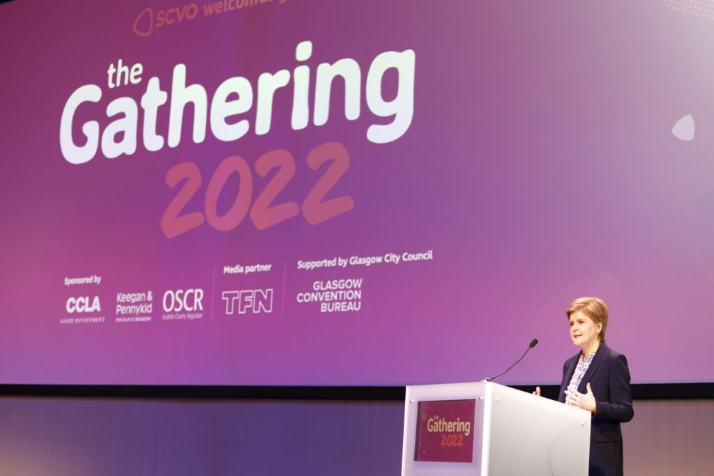 First Minister of Scotland, Nicola Sturgeon MSP stands at a lectern speaking in front of a large screen that says "the Gathering 2022"