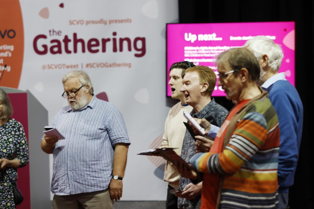 Group of 5 people pictured singing from song sheets in front of signage that says "the Gathering" and a TV secreen that says "Up next..."
