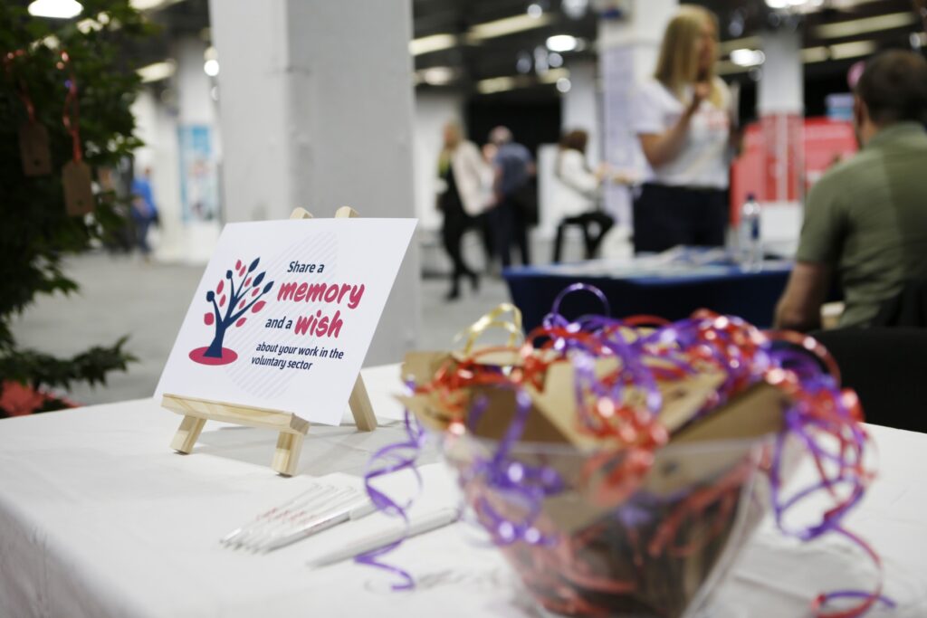 A sign that says "share a memory on a wish" leans on a small wooden easel on top of a table dressed with a white tablecloth. There are pens lying on the table. there is a bowl with brown cards attached to purple and orange ribbon