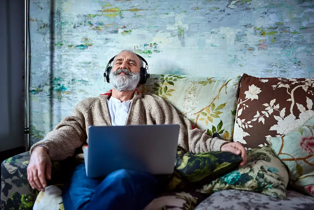 A man sitting on a couch with a laptop, with headphones on.