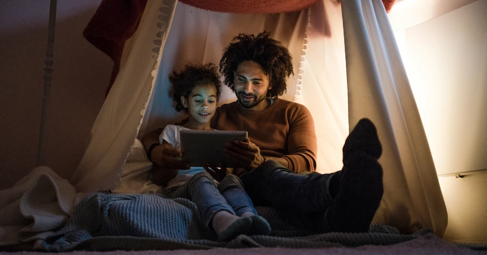 A father and daughter sitting in a pillow fort, looking at a tablet.