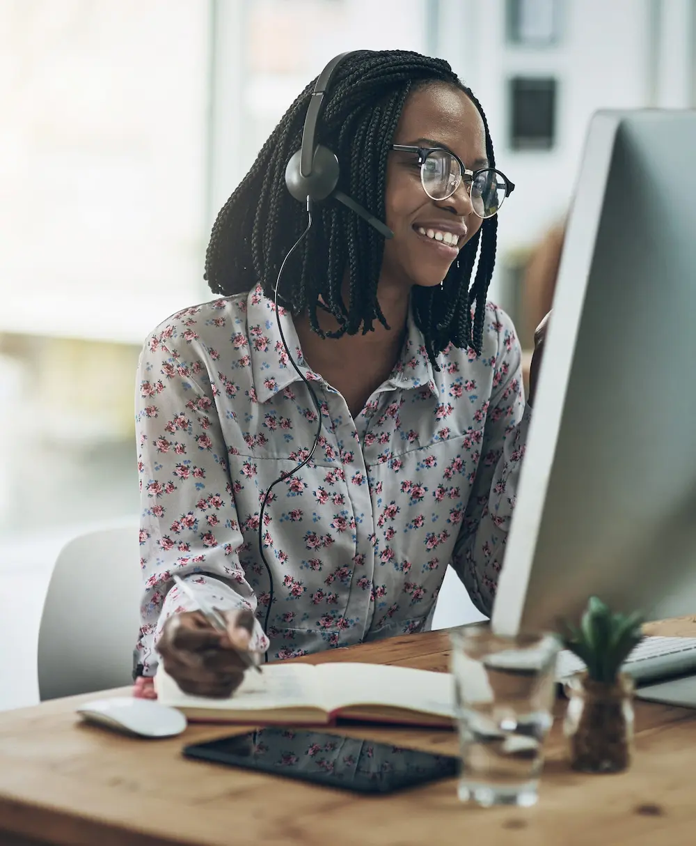 Woman working at desk, taking notes, with a headset on.