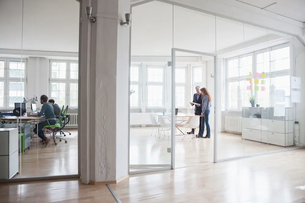 A modern, all white office with glass walls. In one room a man and woman talking in a conference room. In another room two men working at desktop computers.