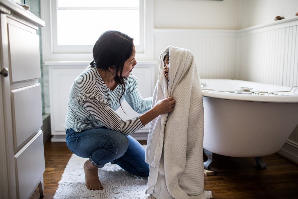 Woman helps small child wrapped fully in a while towel out of bathtub