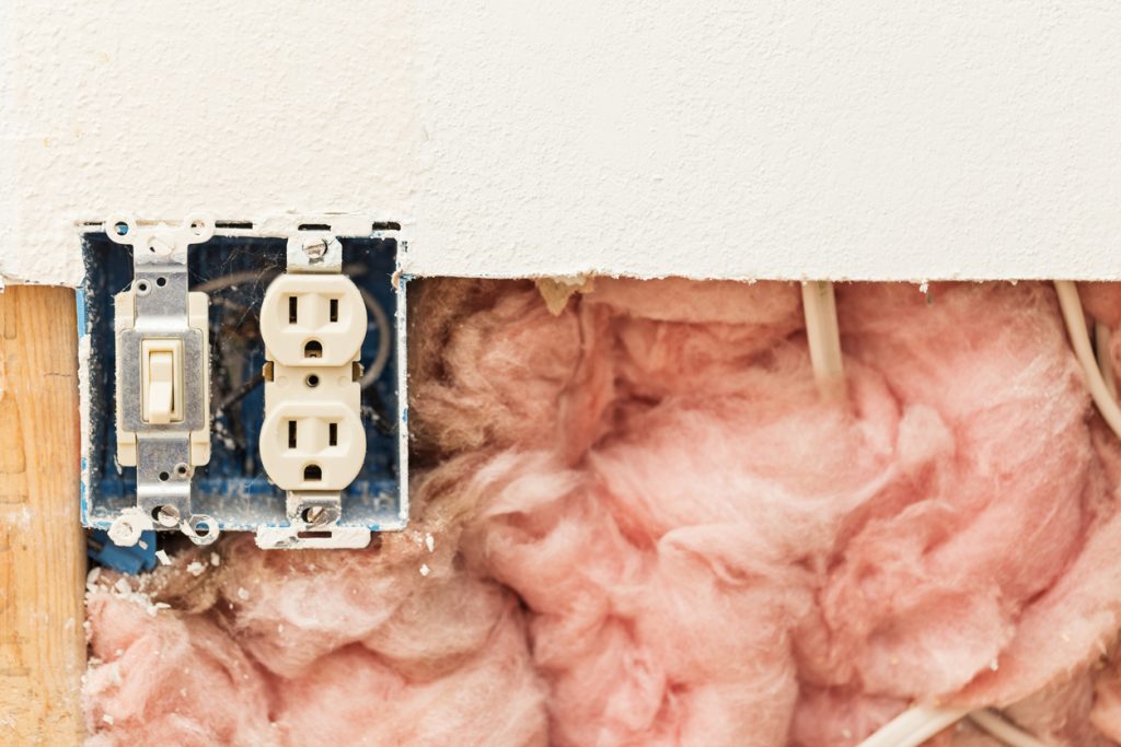 cut out of drywall shows pink fiberglass insulation and electrical wiring with uncovered outlet