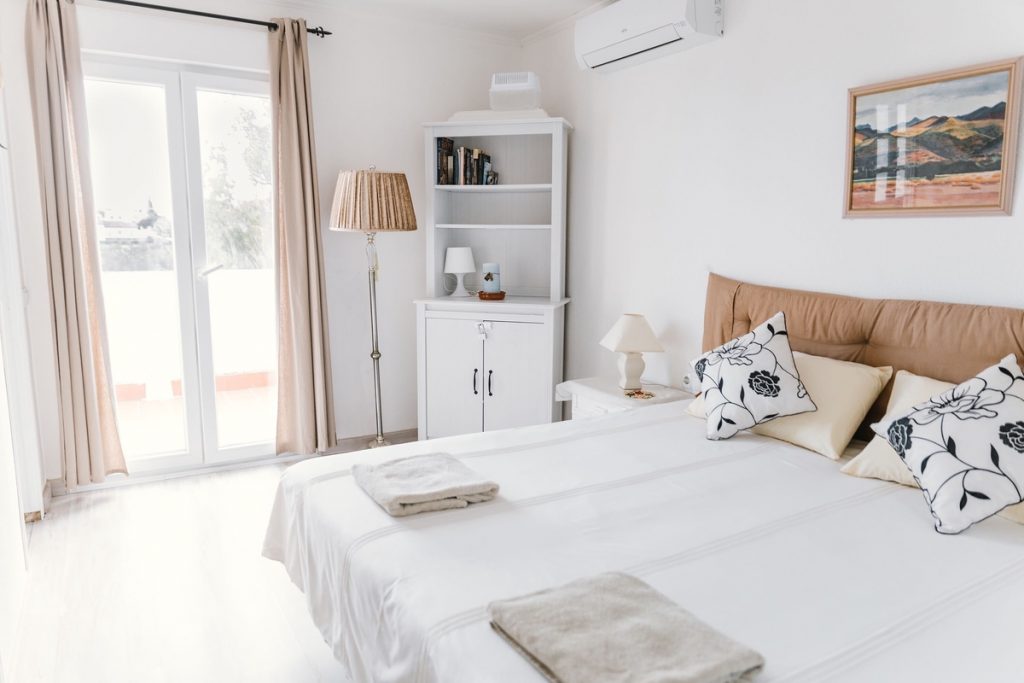 A mini split heat pump unit is featured in a primary, modern bedroom that is well-decorated and well-lit with natural light