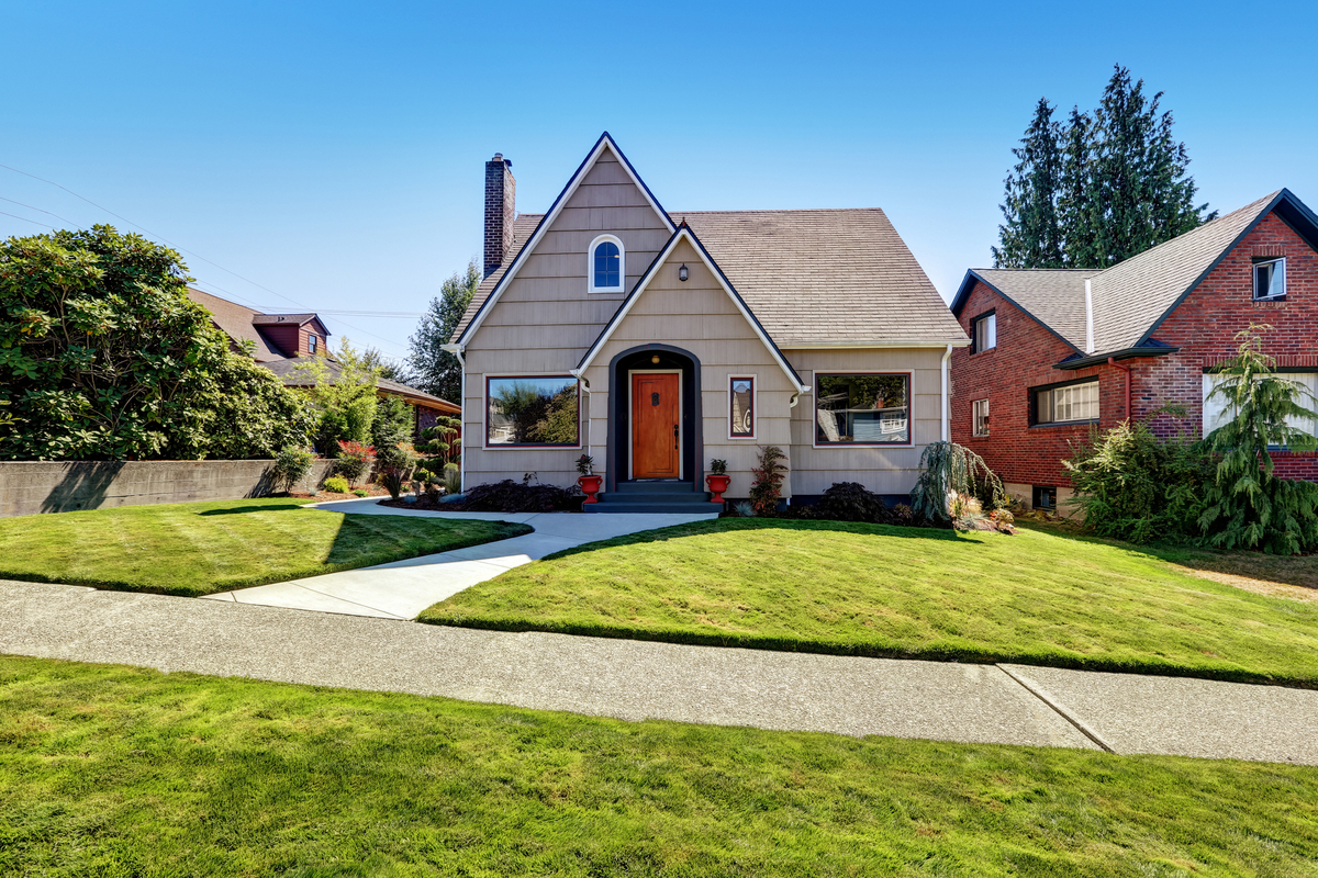 Tan cottage-style house with red door on a beautiful day with perfectly green yard