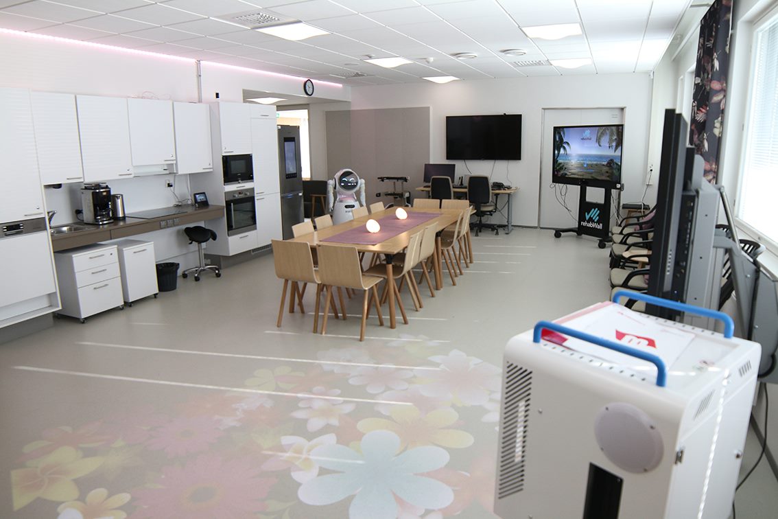 A room with various health-related equipment and TV monitors as well as a kitchenette and a long table with chairs.