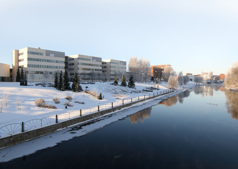 Valkoisia kampusrakennuksia lumen keskellä joen rannalla/ White Campus buildings amidst snow on a river.