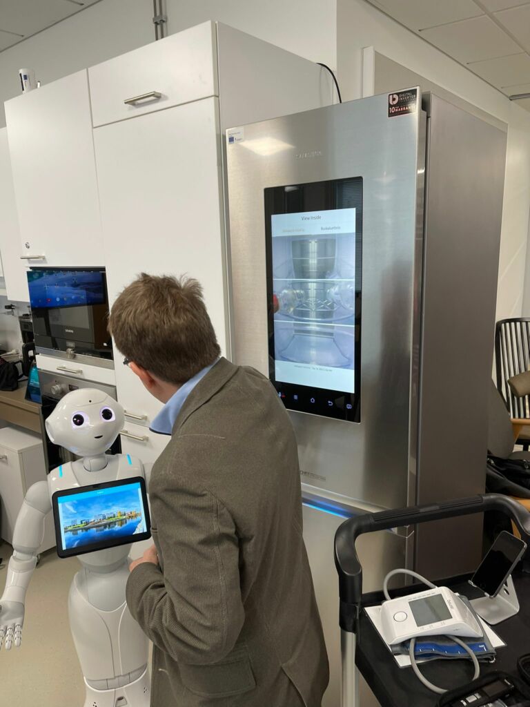 A man and a white robot in front of a metal grey fridge with a touch screen.