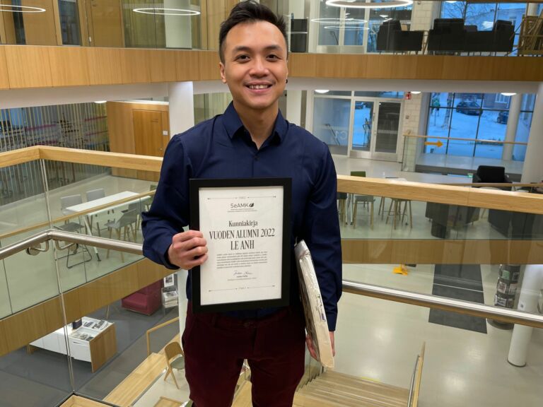 A young man in a blue collared shirt stands with a diploma in his hand.