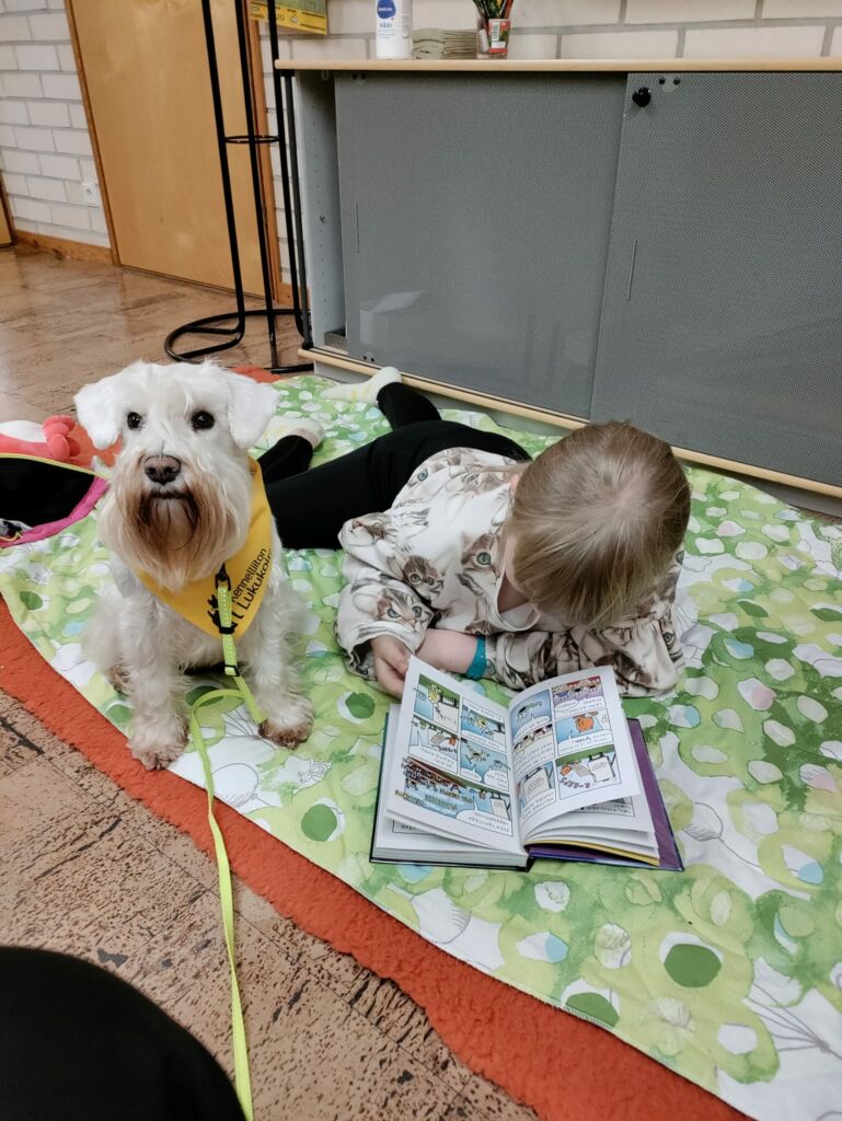 Lapsi lukee kirjaa lattialla, valkoinen, pieni koira istuu vieressä. Koiralla keltainen lukukoira-huivi kaulassa.