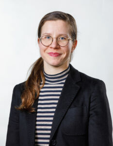 Pitkähiuksinen ja silmälasipäinen henkilö hymuyilee.