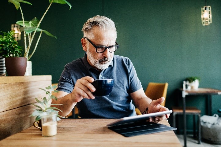 Mies katsoo tablet-tietokonetta pöydän ääressä, kahvikuppi kädessä.