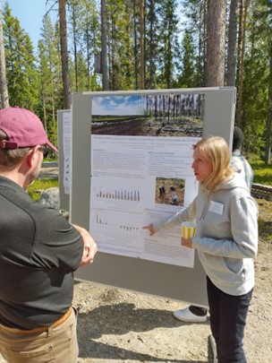 Nainen esittelee posteria ulkona, metsän reunalla.