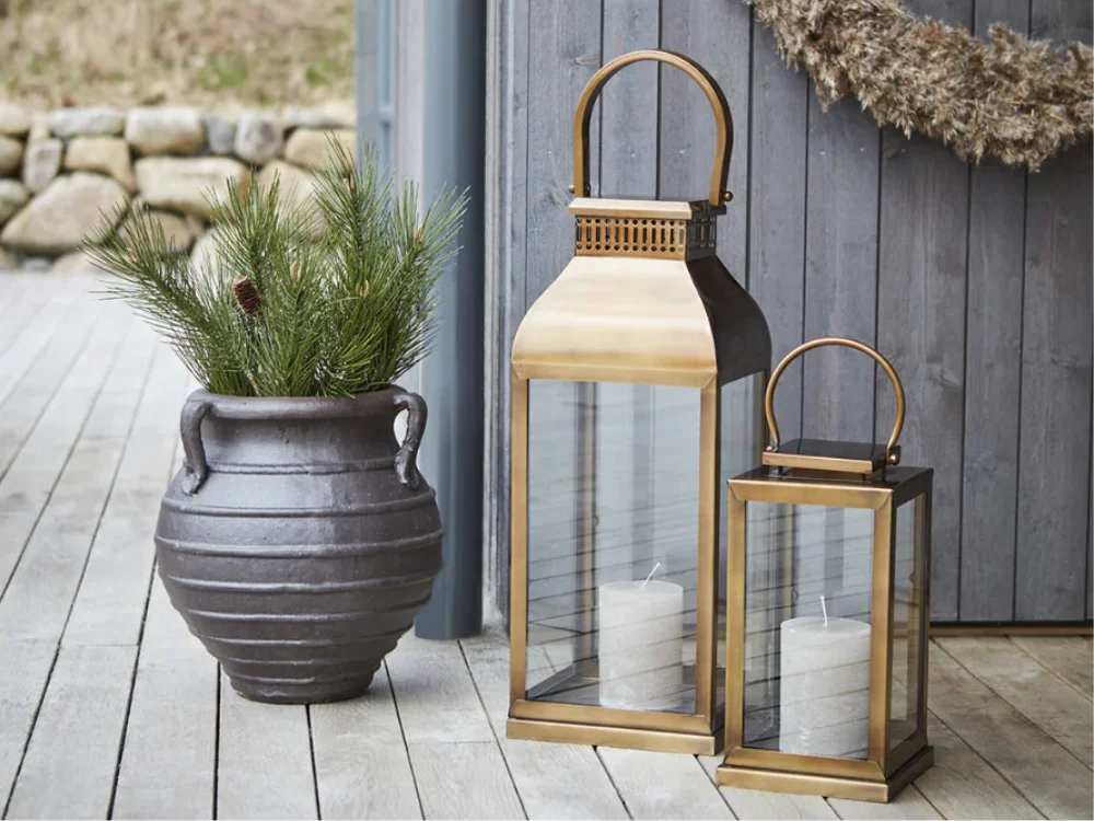 A sophisticated outdoor still life featuring a group of three modern metal lanterns with lit candles next to a large stone planter with green grass on a wooden patio at dusk.