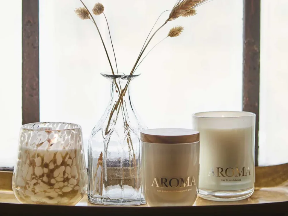 A minimalist scented candle in a dark glass jar sitting on a brass tray atop a marble table, surrounded by warm natural light and soft home textures.