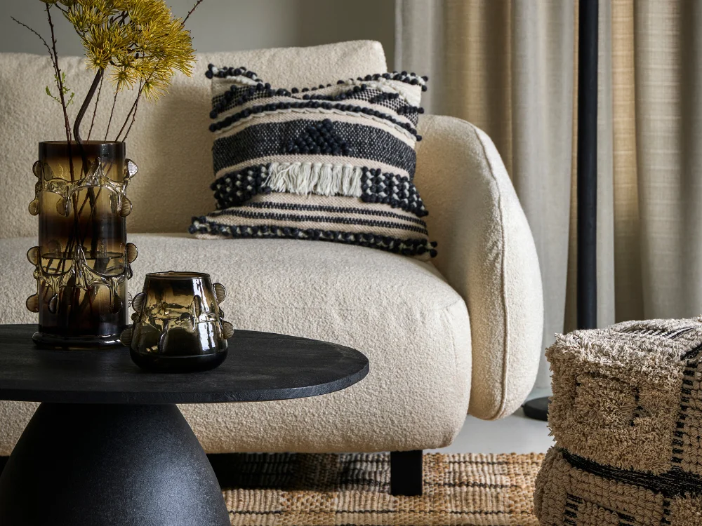 A serene still-life featuring a ceramic vase, a lit scented candle, and a soft wool throw in a room with warm, muted tones.