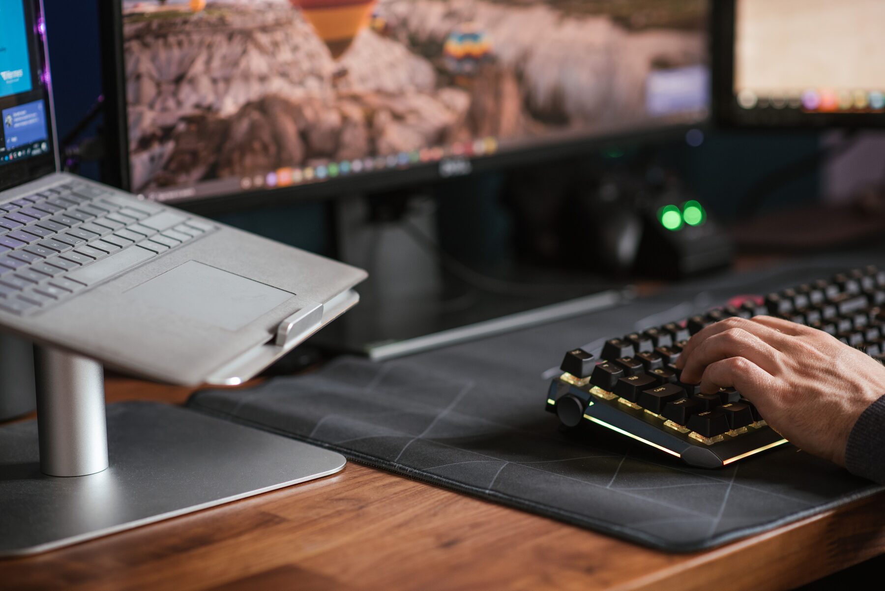Hands typing on a backlit mechanical keyboard beside a laptop raised on a stand