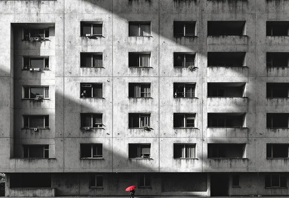 Brutalist concrete housing block at dawn, geometric shadow play, lone red umbre…