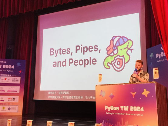 Seth Larson on stage at PyCon Taiwan delivering the keynote "Bytes, Pipes, and People". On the keynote screen there is a cute derpy snake knight character