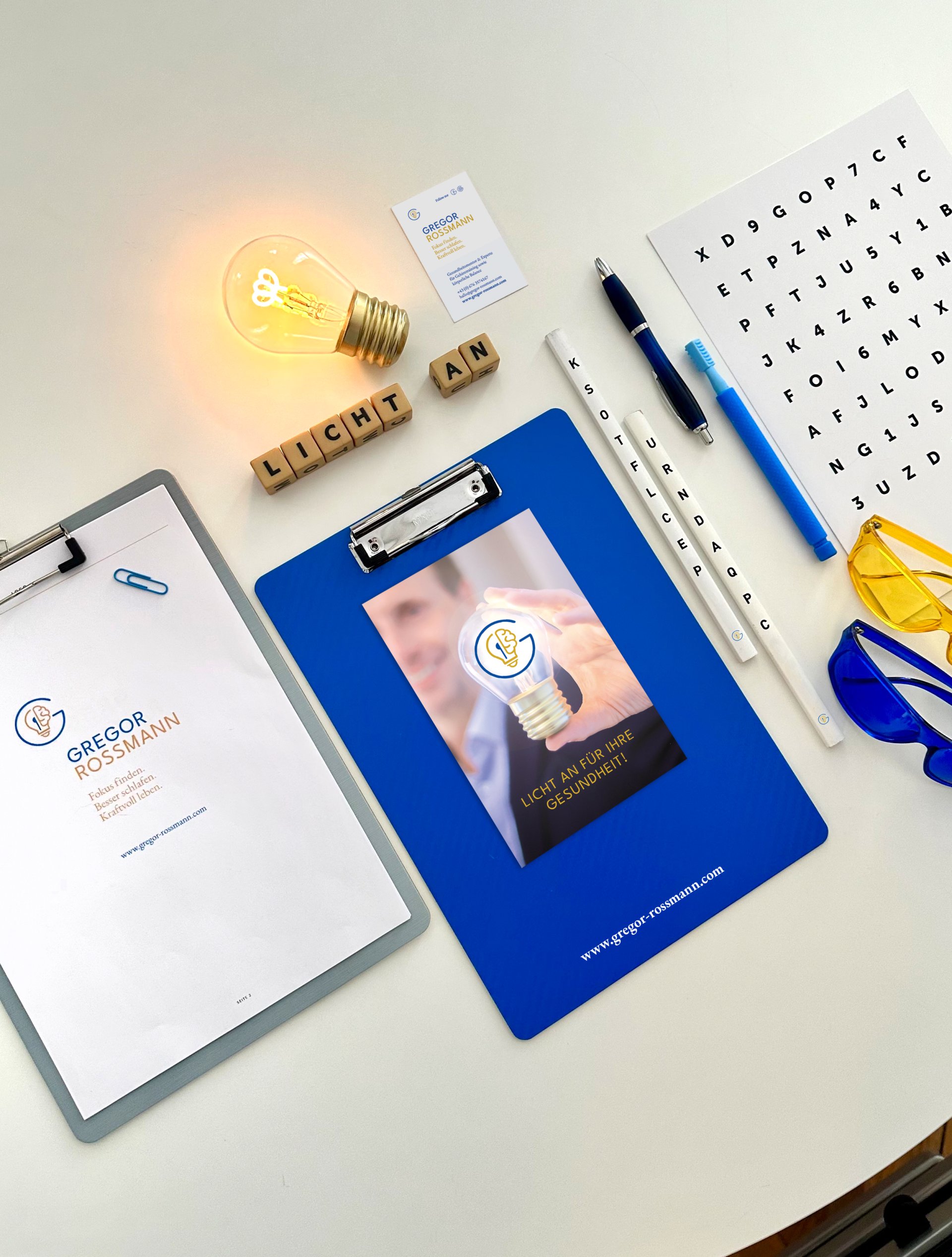 Büroszene mit einem Clipboard, einer Glühbirne, Stiften und einer Visitenkarte von Gregor Rossmann. Das Clipboard zeigt ein ansprechendes Design, während die Glühbirne symbolisch für Ideen steht. Auf dem Tisch liegen auch einige Schreibutensilien und eine Tabelle mit Buchstaben. Die Atmosphäre vermittelt Kreativität und Professionalität.