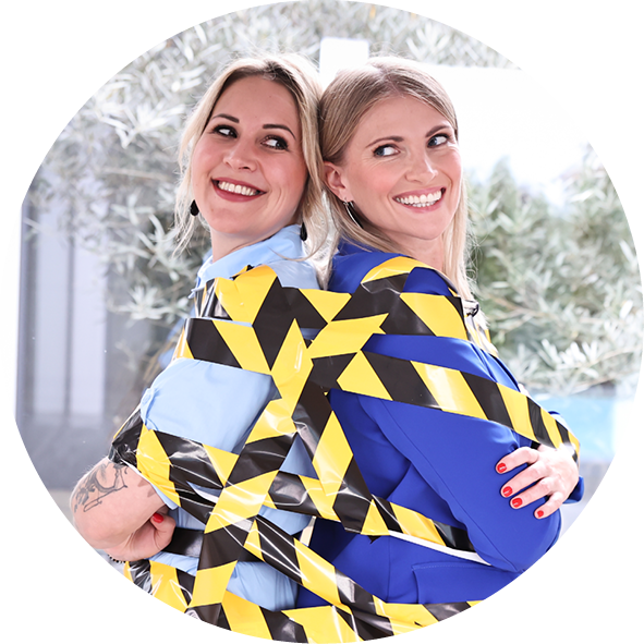 Zwei Frauen stehen Rücken an Rücken, umgeben von gelb-schwarzer Absperrung. Die eine trägt ein hellblaues Hemd, die andere ein blaues Outfit. Beide lächeln freundlich in die Kamera und strahlen Freude aus.