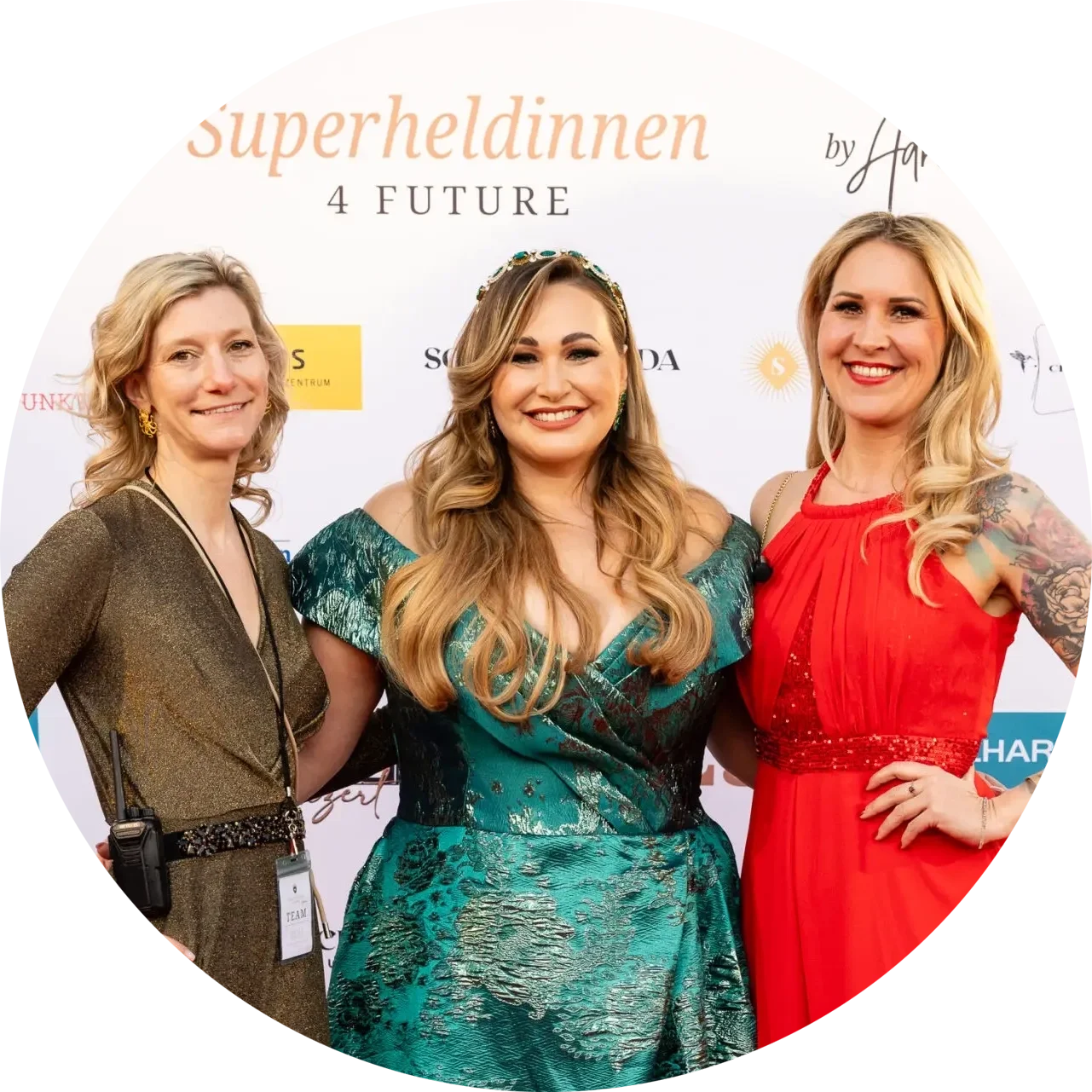 Drei Frauen posieren lächelnd vor einem eleganten Hintergrund bei einer Veranstaltung namens 'Superheldinnen 4 Future'. Die Frau in der Mitte trägt ein auffälliges, glitzerndes Kleid und eine Kopfband, während die anderen beiden in stilvollen Outfits ebenfalls strahlen.