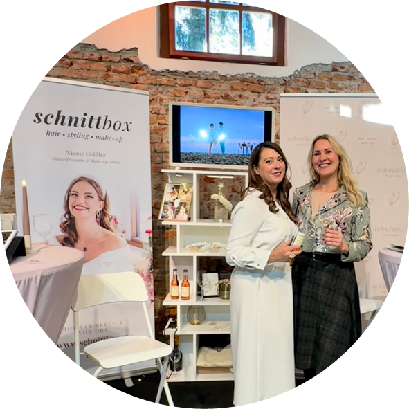 Zwei Frauen stehen lächelnd in einem stilvollen Veranstaltungsraum vor einem Stand der schnittbox, der Dienstleistungen für Haar, Styling und Make-up anbietet. Im Hintergrund ist ein Bildschirm mit einer Strandaufnahme zu sehen. Die Frauen halten Gläser in der Hand und tragen elegante Outfits. Der Stand präsentiert eine einladende Atmosphäre für Hochzeits- und Event-Dienstleistungen.
