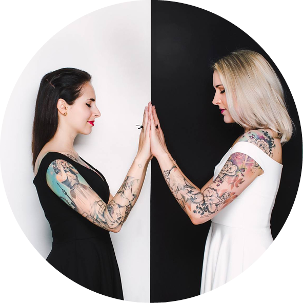 Zwei Frauen mit auffälligen Tattoos stehen sich gegenüber und berühren sich mit den Händen. Die linke Frau trägt ein schwarzes Kleid, während die rechte Frau ein weißes Kleid anhat. Der Hintergrund ist in zwei Hälften geteilt – eine Seite schwarz und die andere weiß. Diese harmonische Darstellung symbolisiert Kontraste und Einheit.