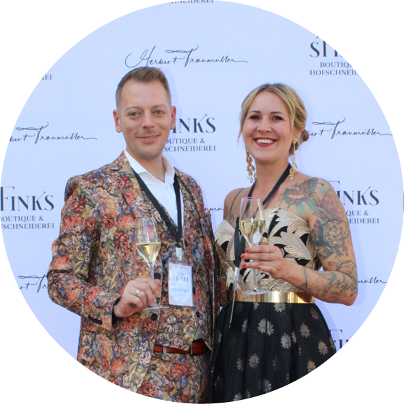 Ein Paar, das an einem festlichen Event teilnimmt, hält Champagnergläser in der Hand. Der Mann trägt einen bunten Anzug mit floralem Muster, während die Frau ein elegantes schwarzes Kleid mit goldenem Gürtel und Tattoos trägt. Im Hintergrund ist ein Banner mit dem Schriftzug "Finks Boutique & Hofschneiderei" zu sehen.