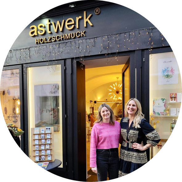 Zwei Frauen stehen lächelnd vor dem Geschäft 'astwerk' für Holzschmuck. Das Schaufenster zeigt eine einladende Auswahl an handgefertigtem Schmuck und dekorativen Elementen. Die Atmosphäre ist freundlich und einladend, perfekt für Schmuckliebhaber.
