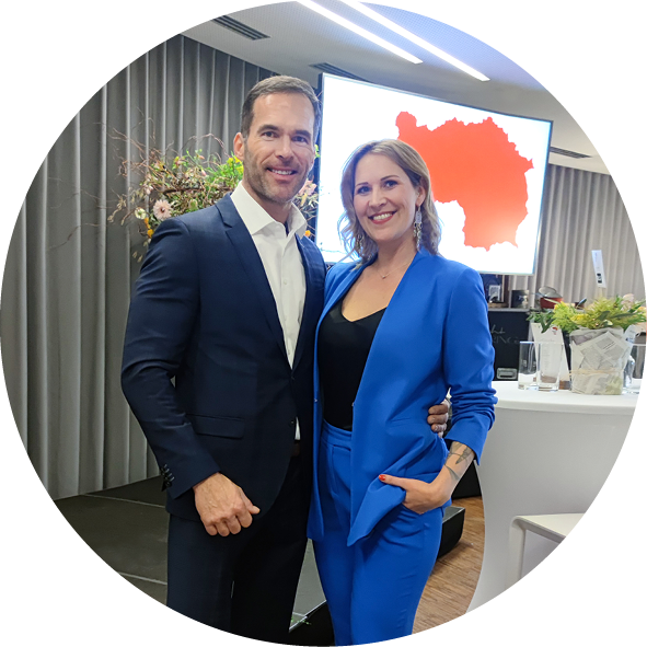 Ein Paar posiert freundlich für ein Foto auf einer Veranstaltung. Der Mann trägt einen eleganten Anzug in Dunkelblau und ein weißes Hemd, während die Frau einen stilvollen blauen Anzug mit einem schwarzen Oberteil kombiniert. Im Hintergrund ist eine Bildschirmpräsentation sichtbar, die eine rote Karte zeigt.