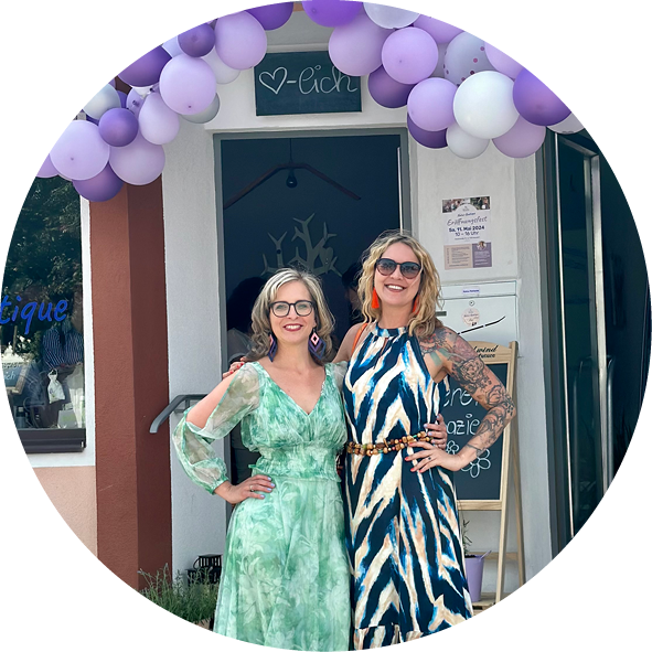 Zwei Frauen stehen vor einem Boutique-Eingang, umgeben von lila Luftballons. Die Frau links trägt ein grünes, gemustertes Kleid und die Frau rechts ein gestreiftes Kleid in Blau- und Erdtönen. Beide lächeln freundlich in die Kamera, während sie die Eröffnung eines neuen Geschäfts feiern.