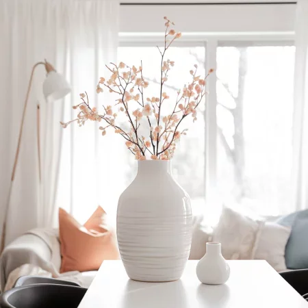 Ein eleganter, weißer Vase mit zarten Blütenzweigen steht auf einem Tisch in einem hellen Raum. Im Hintergrund sind ein Sofa mit Kissen in sanften Farben und große Fenster zu sehen, die Tageslicht hereinlassen.