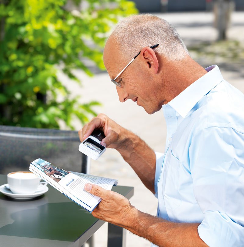 Älterer Mann mit Brille nutzt Lupe mobilux LED, um in einem Buch zu lesen, sitzt draußen an einem Tisch mit einer Tasse Kaffee