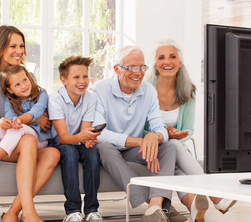 Mehrgenerationenfamilie sitzt auf Sofa und schaut gemeinsam fern, älterer Mann trägt TV-Brille maxTV