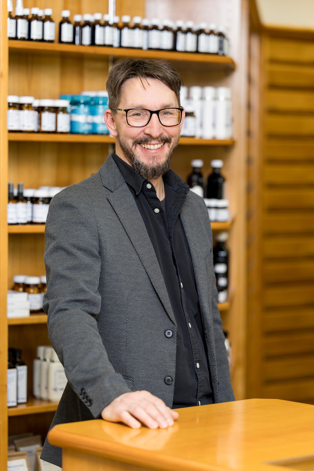 Porträt eines Mannes mit Brille und Bart, der an einem Empfangstresen in einem Geschäft steht. Im Hintergrund sind Regale mit verschiedenen Flaschen und Behältern zu sehen, die eine Vielzahl von Produkten enthalten. Der Mann trägt einen grauen Blazer über einem schwarzen Hemd.
