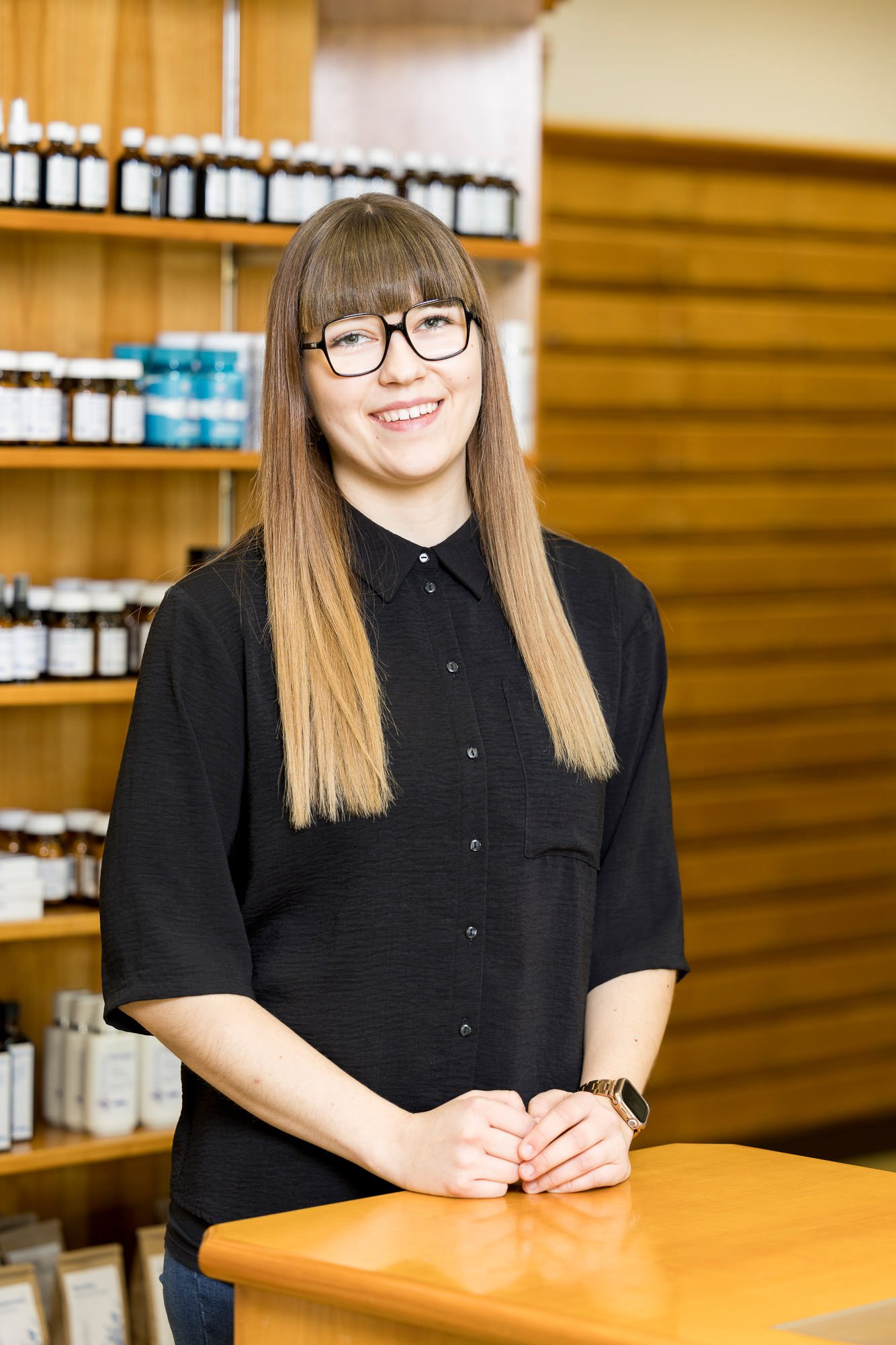Eine junge Frau mit langen, glatten Haaren und einer Brille steht an einem Empfangstresen in einem Geschäft. Sie trägt ein schwarzes Hemd und lächelt, während im Hintergrund Regale mit verschiedenen Produkten sichtbar sind.