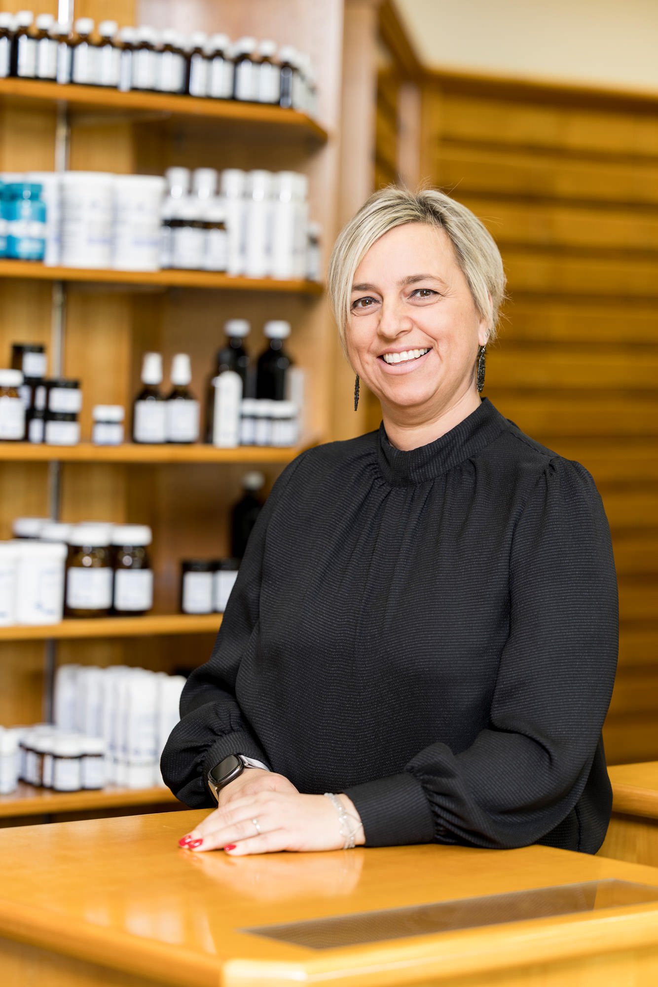 Eine lächelnde Frau mit blonden Haaren steht an einem Empfangstresen in einem Geschäft. Im Hintergrund sind Regale mit verschiedenen Flaschen und Produkten sichtbar, die für eine Apotheke oder ein Wellness-Center typisch sind. Sie trägt ein schwarzes Oberteil und hat ihre Hände auf dem Tisch platziert.